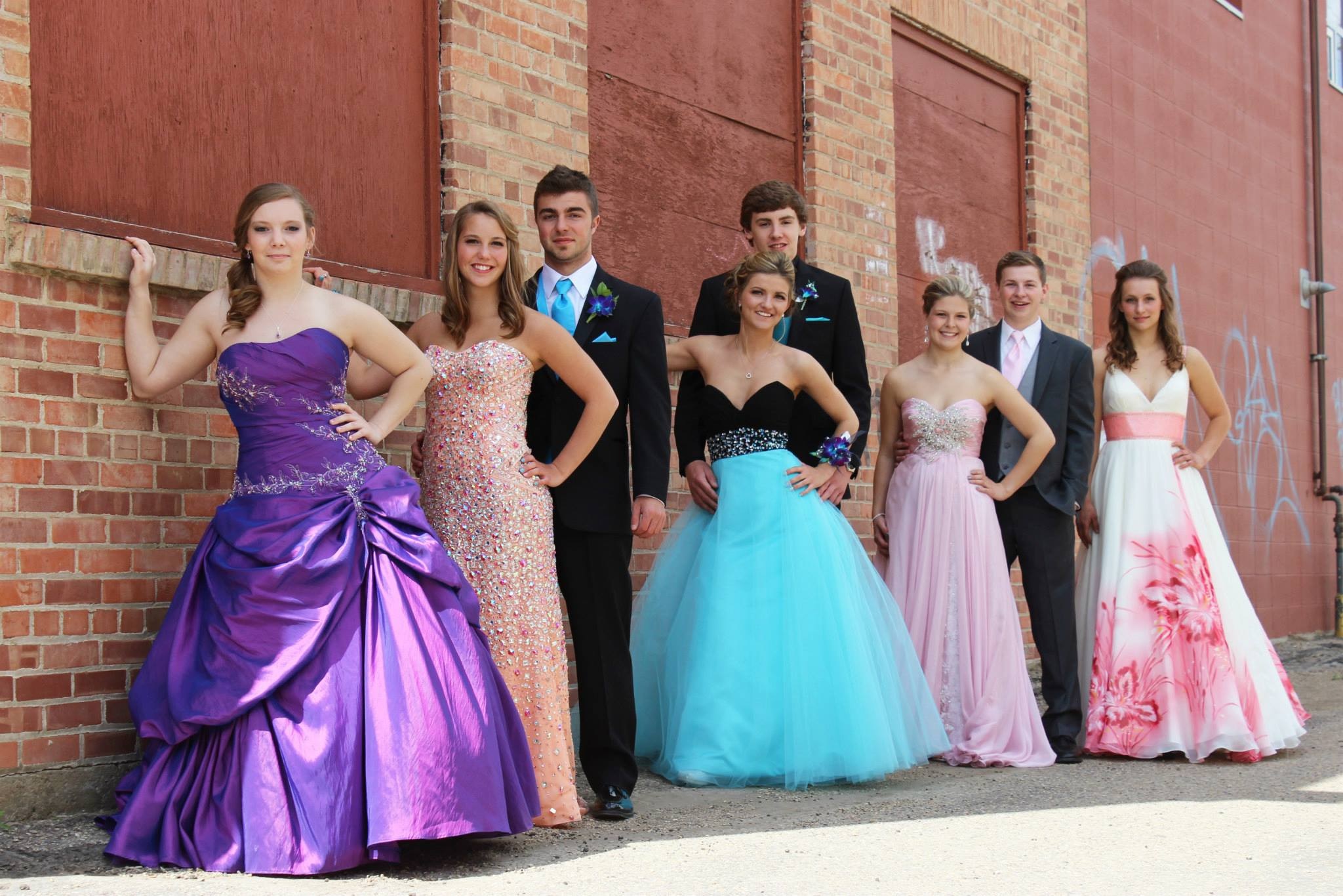 Prom Photographer A group of female and male graduates stand posed in a line against a brick wall, smiling at the camera