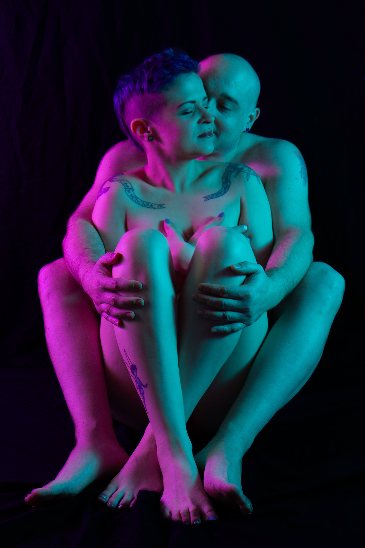 Couples Boudoir A woman sits in front of and between her husbands legs. He cradles her with his arms around her. She turns her head back for a kiss on the cheek. Split lighting with pink and teal light.