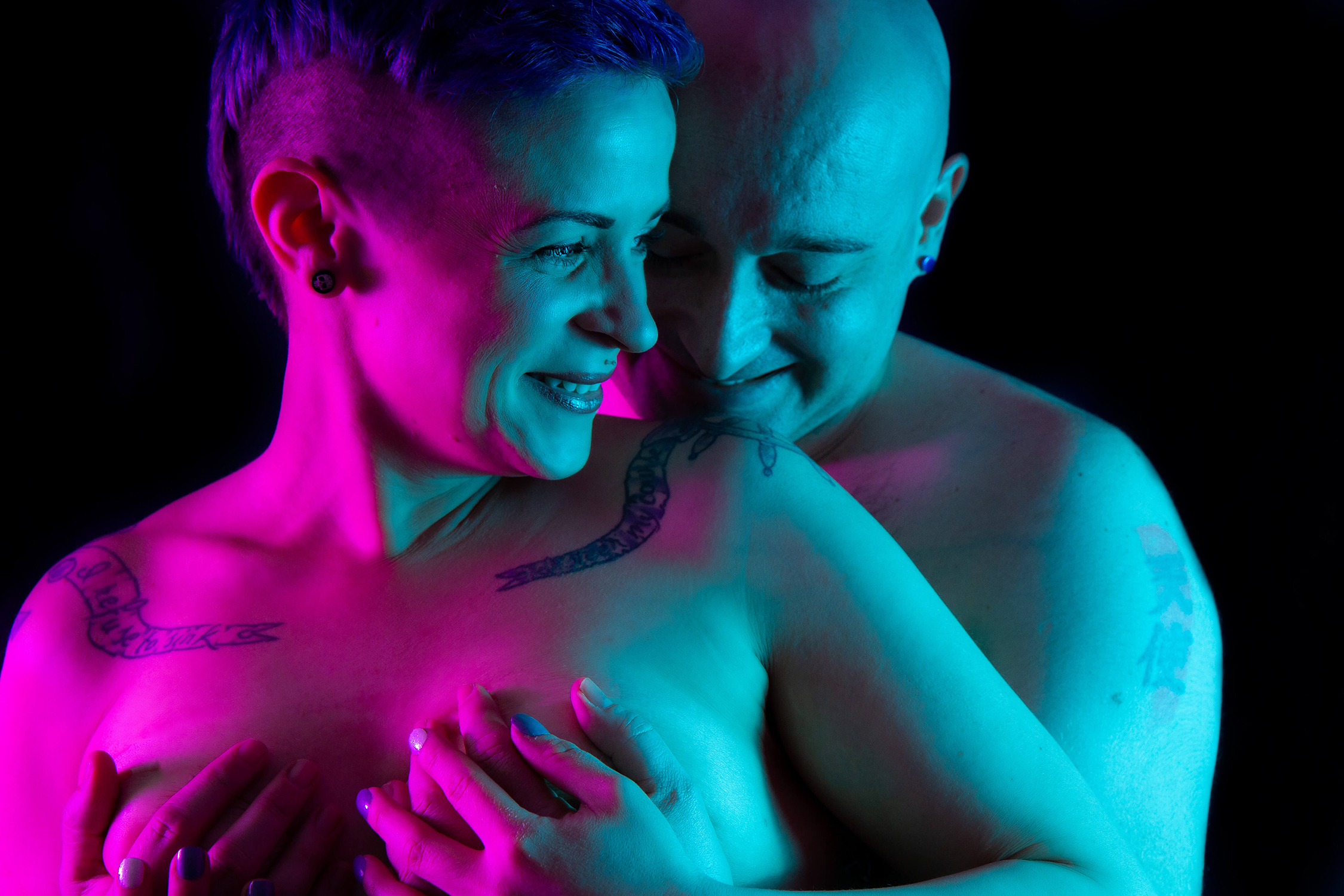 Couples Boudoir a man hugs a woman from behind as they smile at each other. Split lighting with pink and teal light.