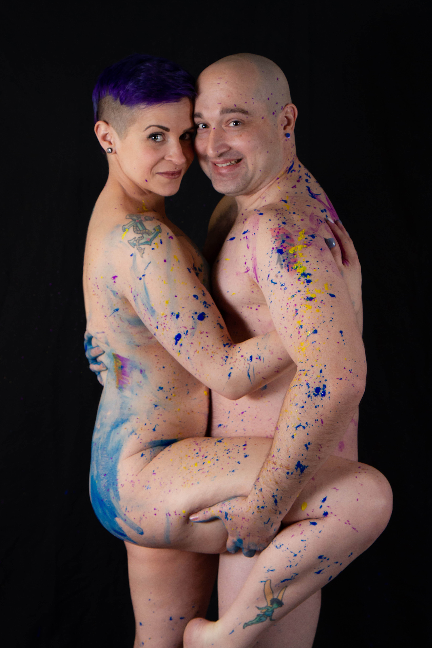 Couples Boudoir a man and a woman, nude, are covered in paint spatter, embracing chest to chest, arms around one another, smiling at the camera